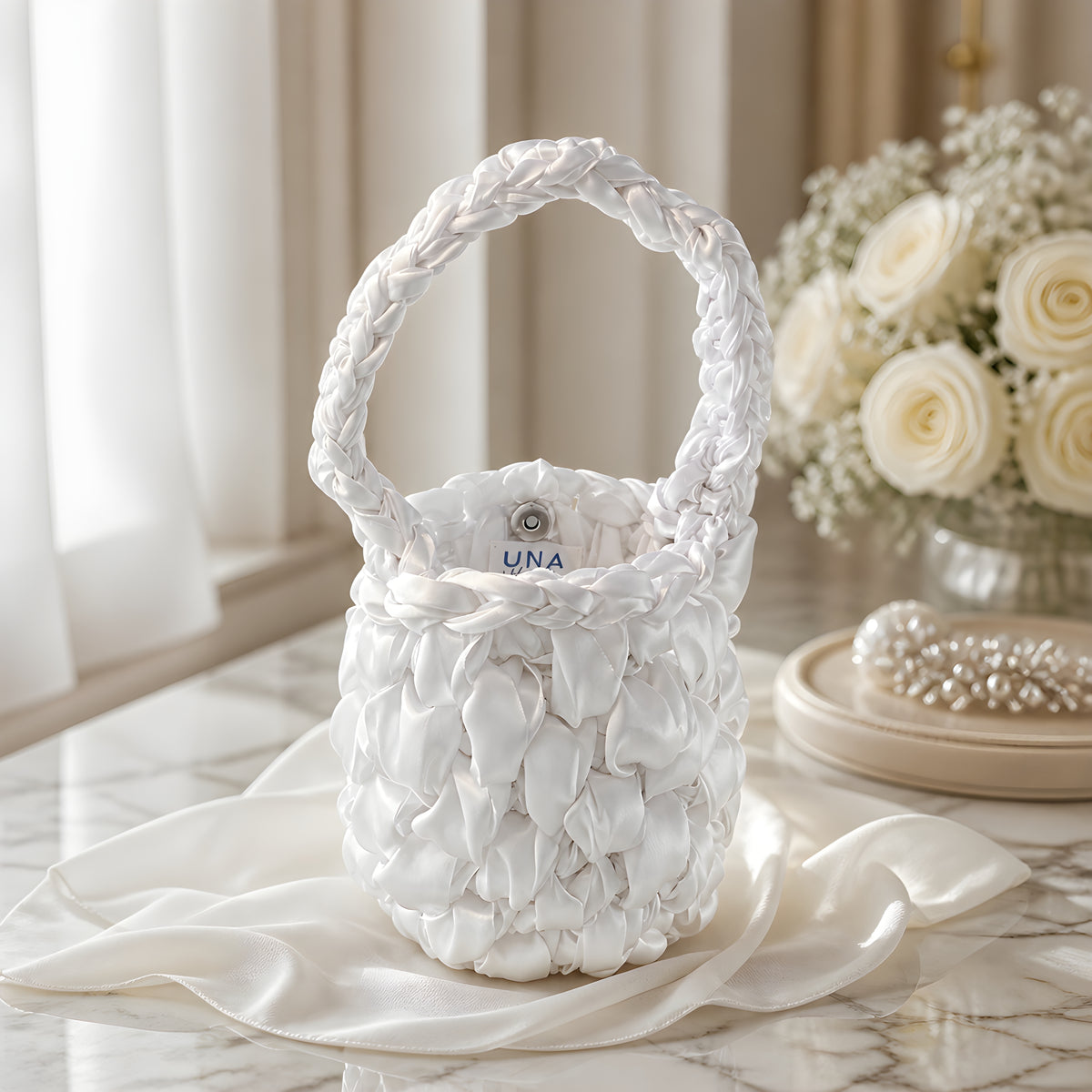 White satin crochet handbag with 'UNA' branding on a marble surface with a floral arrangement in the background.